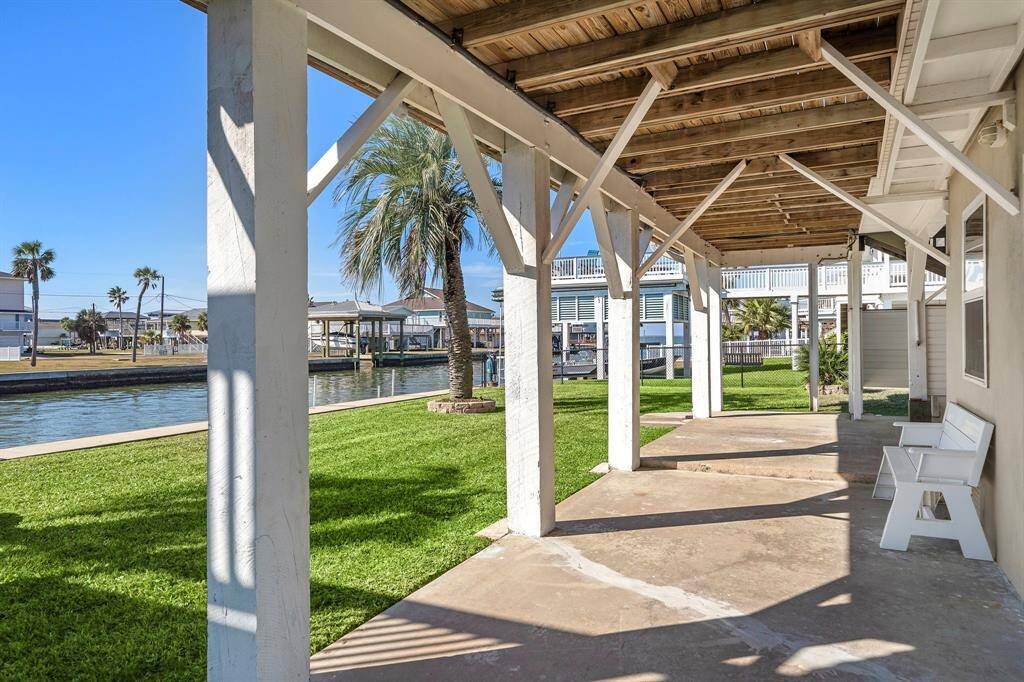 A view along the back of the home from beneath the deck, showing the wide lawn and water frontage. Palm trees and the bulkhead line the canal, reinforcing the coastal lifestyle and easy water access.