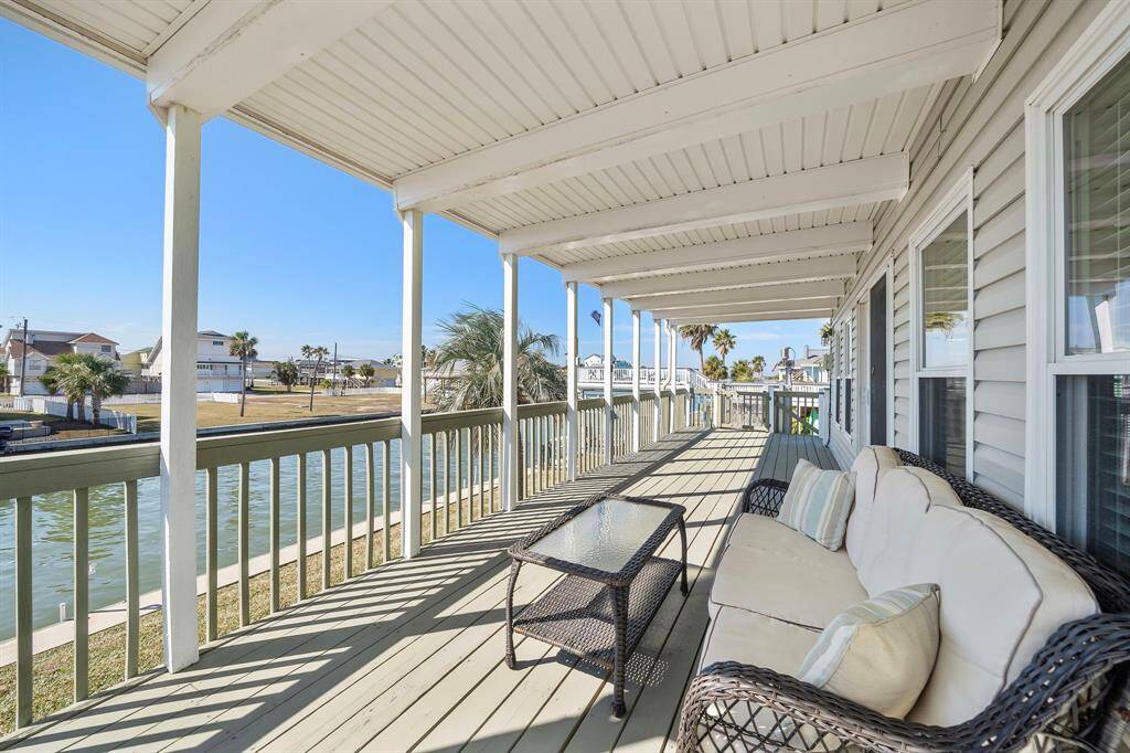 A covered waterfront deck with a plush outdoor sofa and glass coffee table creates an inviting space to relax while enjoying serene canal views. The extended roof provides shade, making it the perfect spot for lounging at any time of day.