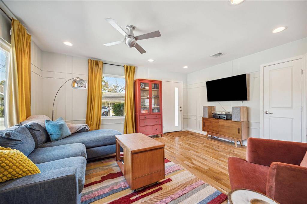 The warm and welcoming living room features an open, bright design with modern wall paneling, MoMa ceiling fan, recessed lighting, and large windows providing tons of natural light.