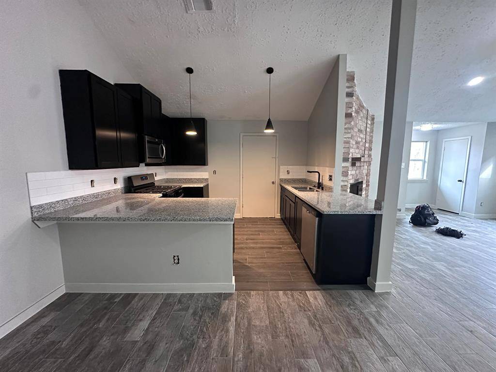 This photo showcases a modern kitchen with granite countertops, dark cabinetry, and stainless steel appliances. It features an open layout with pendant lighting and connects to a spacious living area with a visible brick accent wall. The room has a sleek, contemporary design with wood-look flooring.