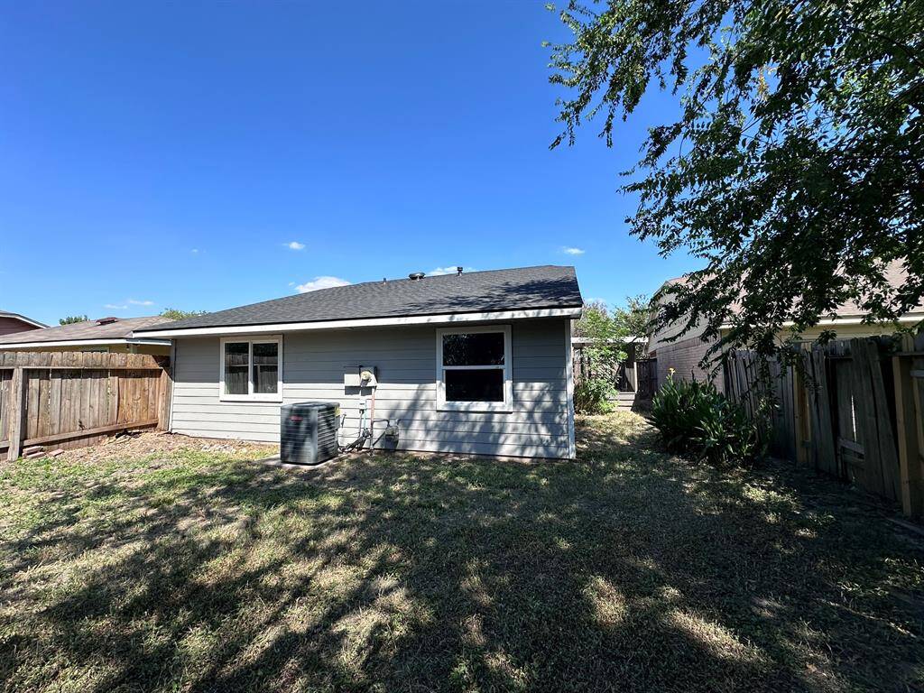 This photo shows the backyard of a home with a small lawn area, wooden fencing, and an exterior view of the house's back, featuring a window and an air conditioning unit. The yard is partially shaded by a tree, providing a cozy outdoor space.
