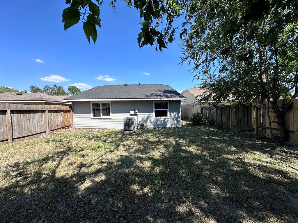 This photo shows a modest backyard with a grassy area, surrounded by wooden fencing. The back of a small, single-story house is visible, featuring a window and an air conditioning unit. The yard has a few trees providing shade and a clear blue sky above.