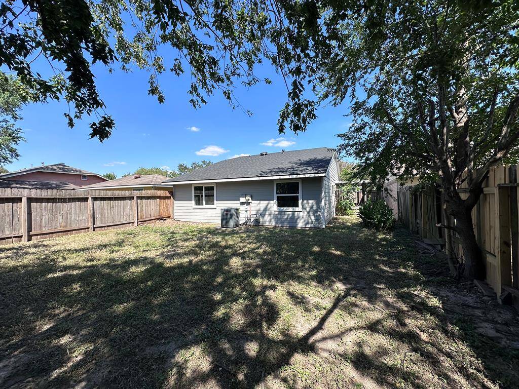 This is a backyard view of a single-story house with a fenced yard. It features a grassy area with some trees, offering shade and privacy. The exterior is painted light gray, and there is an air conditioning unit visible on the side.
