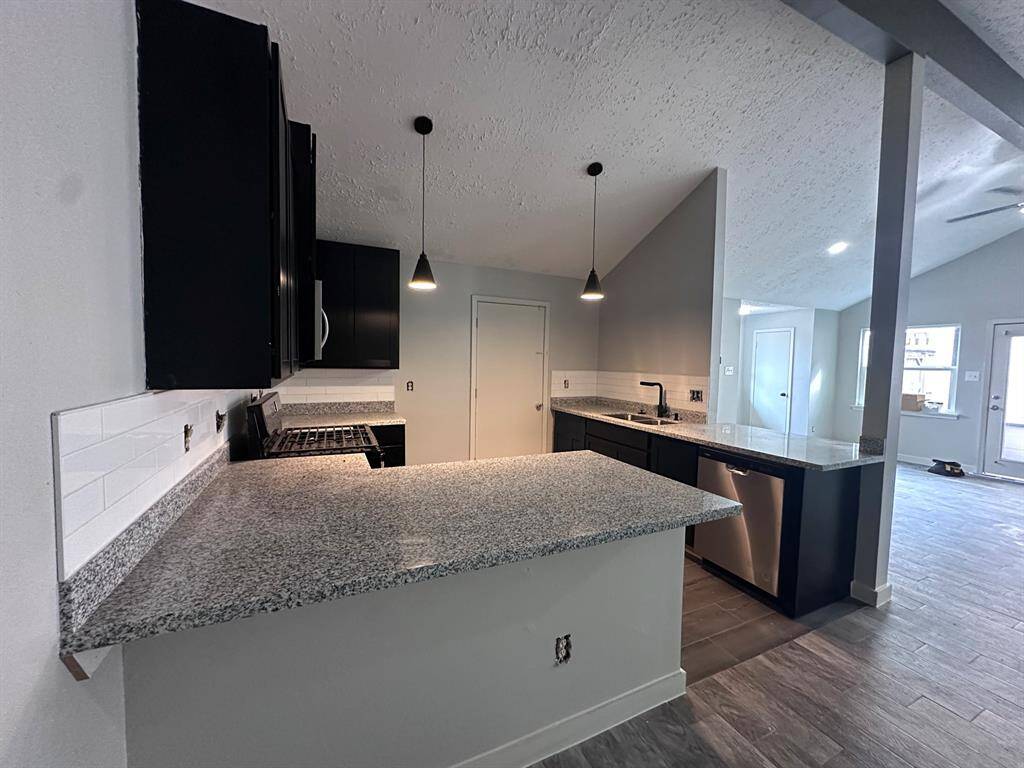 Modern kitchen with granite countertops, dark cabinetry, and stainless steel appliances. Features a peninsula with pendant lighting and an open layout leading to a bright living area with large windows.