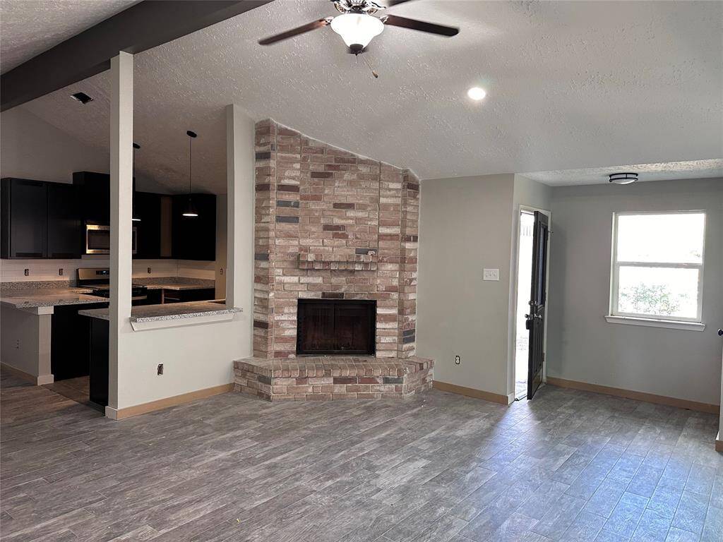 This photo showcases a modern living area with a vaulted ceiling, featuring a stylish brick fireplace. The space is open, connecting to a kitchen with dark cabinetry and granite countertops. Natural light enters through a window and door, highlighting the wood-look tile flooring.