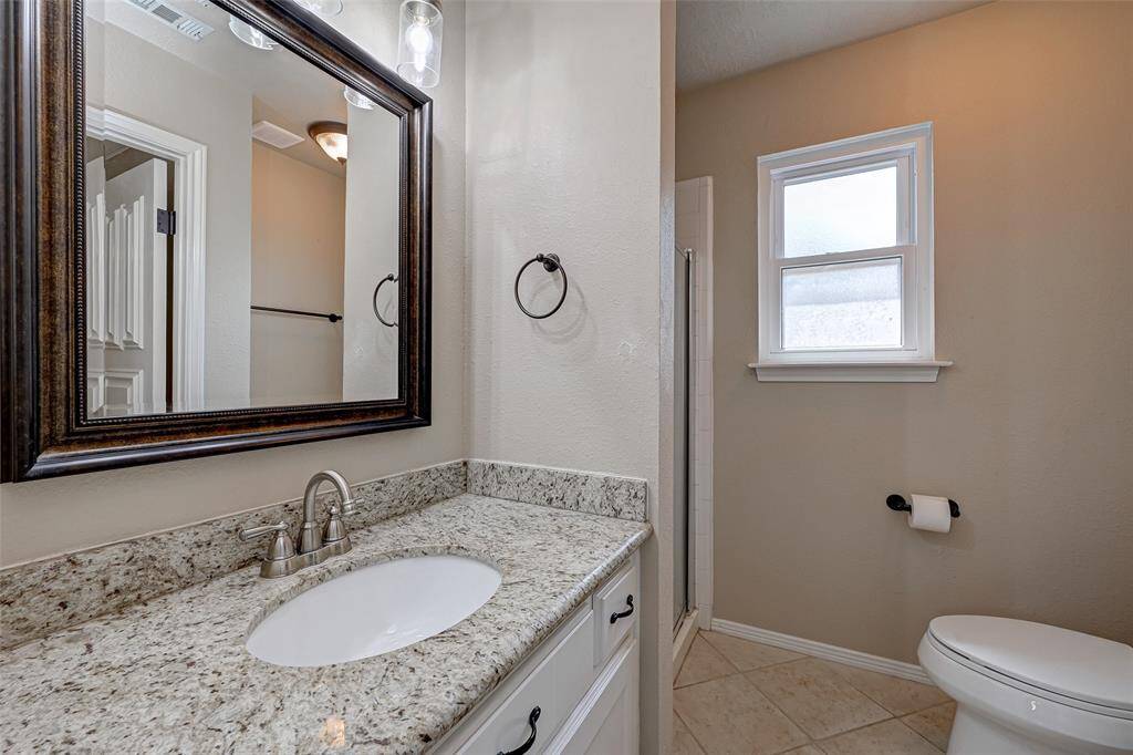 Bathroom #3 is well-appointed with a single sink set in a GRANITE vanity, accompanied by a decorative framed mirror and updated lighting for a stylish touch. It also boasts a TILED WALK-IN SHOWER with a glass door and includes a commode, offering both functionality and modern aesthetics.