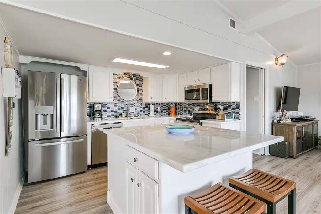 This modern kitchen features stainless steel appliances, a large island with seating, and sleek white cabinetry. The stylish backsplash adds a touch of elegance, and the open layout flows into the living area with a mounted TV.