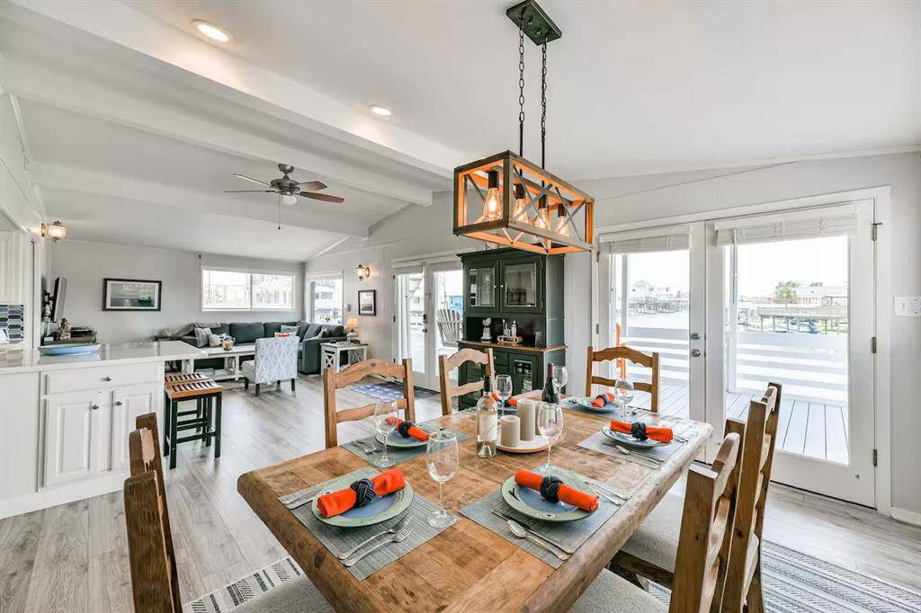 This inviting open-concept living space features a rustic dining area with a wooden table set for six, a cozy living room with ample seating, and large windows and doors leading to a deck with waterfront views. The room is bright and airy, with modern light fixtures and neutral tones creating a warm atmosphere.