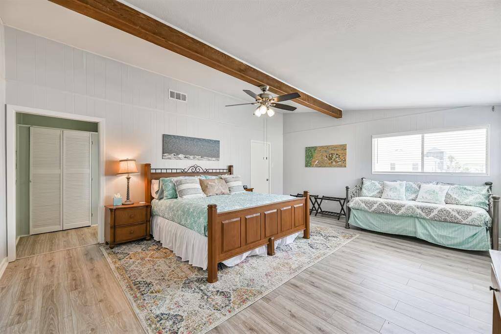 Spacious bedroom with light wood flooring, featuring a wooden bed, a ceiling fan, and a large window for natural light. A cozy sitting area with a daybed is also included, and the room has a warm, inviting atmosphere.