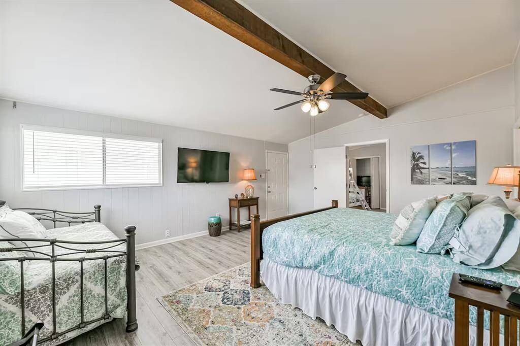 This spacious bedroom features a vaulted ceiling with an exposed beam, a ceiling fan, and ample natural light from a large window. It includes a cozy bed, a daybed, a wall-mounted TV, and tasteful decor, creating a comfortable and inviting atmosphere.