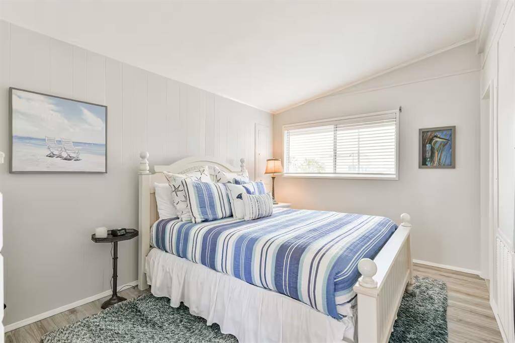 This bright, cozy bedroom features a striped blue and white bedspread, a window with blinds for natural light, and tasteful beach-themed artwork. The room has light-colored walls and flooring, creating a serene, inviting atmosphere.