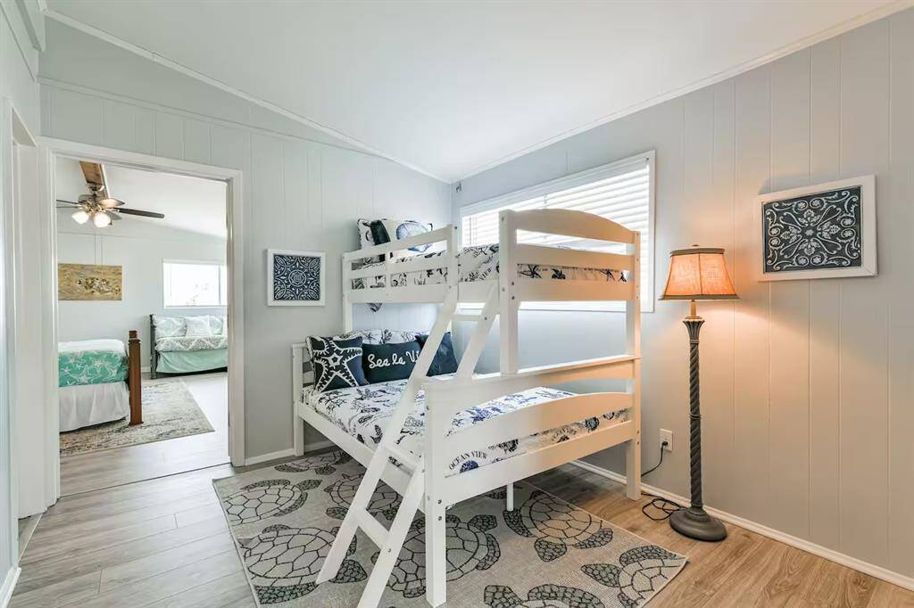 This cozy bedroom features a bunk bed with nautical-themed bedding, light gray walls, and wood flooring. A soft rug adds warmth.  An adjacent room with a ceiling fan is visible, enhancing the open feel.