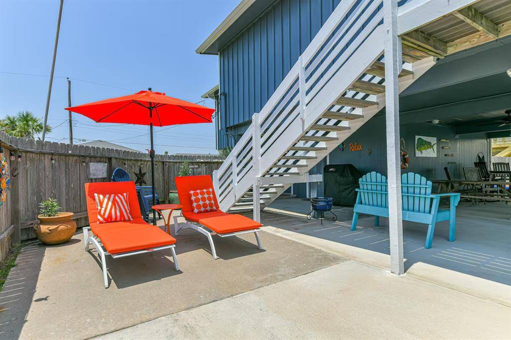 This inviting outdoor space features a concrete patio with vibrant orange loungers and a matching umbrella, perfect for relaxation. A covered area with seating and a grill offers shade and entertainment options. The wooden fence provides privacy, enhancing the cozy atmosphere.