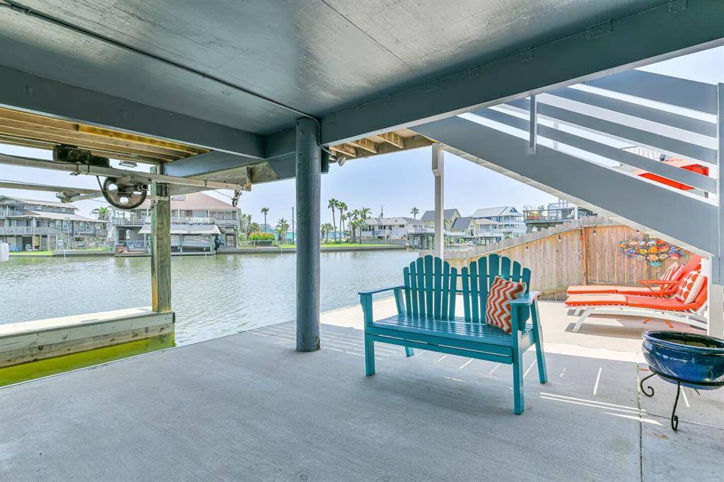 This waterfront patio features a covered seating area with a turquoise bench and colorful cushions, offering scenic views of a canal. There's a staircase leading up to the sun deck/crow's nest, a grill, and additional lounging space with vibrant decor, ideal for relaxation and entertaining.