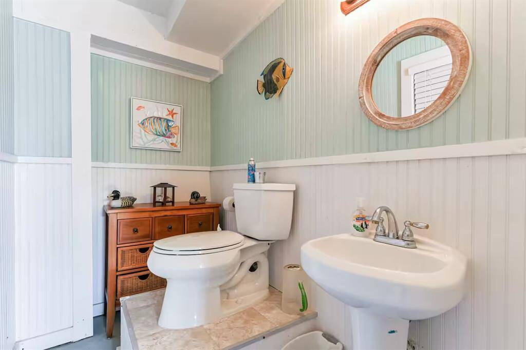 This bathroom features a cozy coastal theme with light green and white paneling. It includes a toilet, a pedestal sink, a round mirror, and decorative fish art, creating a fresh, inviting space.