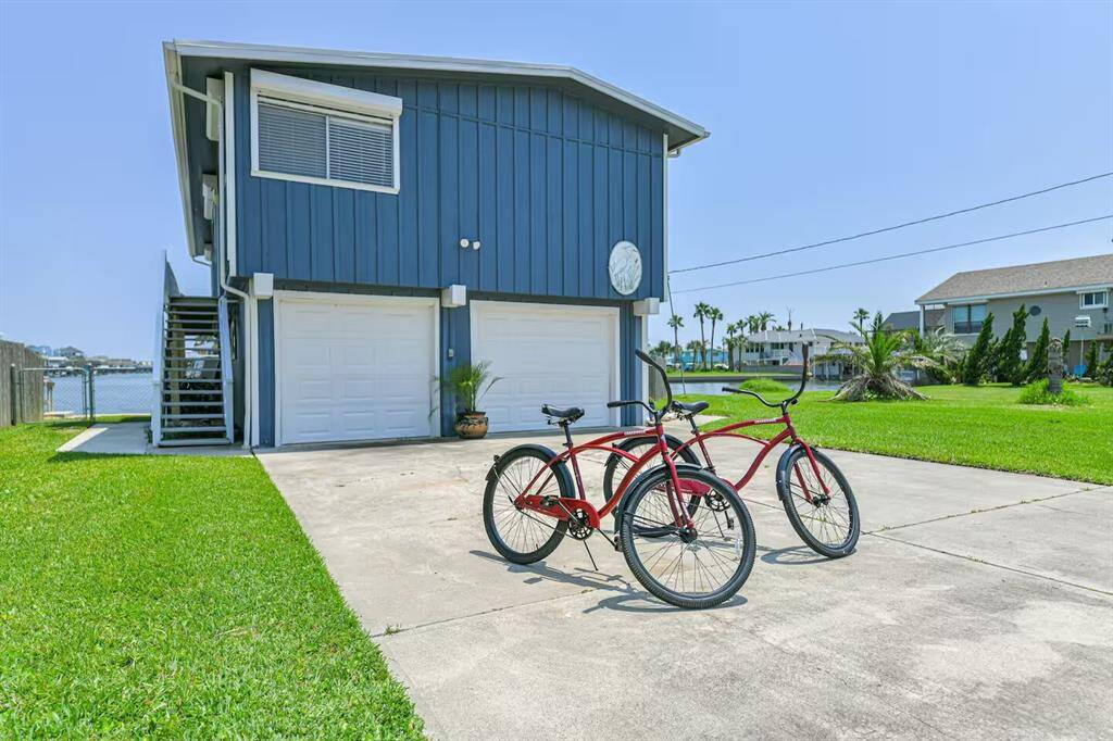 This charming one-story coastal home features a spacious driveway with two bicycles, a blue exterior, and a double garage. It's located in a serene neighborhood with water views and lush greenery.