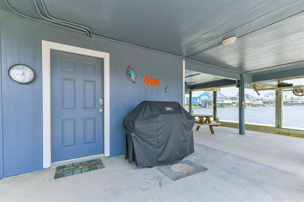 This photo showcases a covered patio area with a blue door, wall clock, and decorative elements. There's a gas barbecue grill, picnic table, and views of the waterfront, offering a serene outdoor space perfect for relaxation and entertaining.
