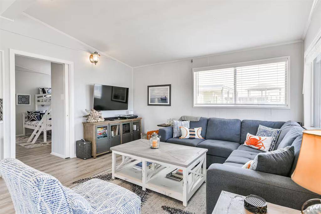 This inviting living room features a cozy L-shaped sofa, a large window with abundant natural light, and a wall-mounted TV. The decor is coastal-inspired with nautical accents, creating a relaxed atmosphere. Adjacent is a glimpse into a room with bunk beds, suggesting a family-friendly layout.