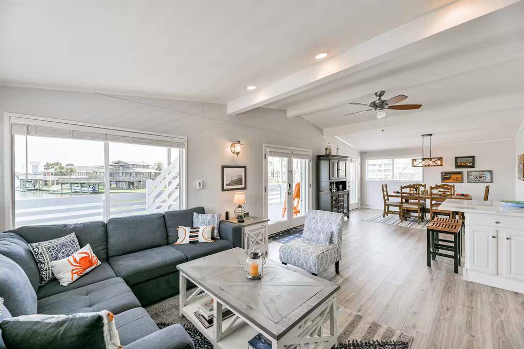 This bright, open-concept living space features large windows, a comfortable sectional sofa, and modern decor. It seamlessly connects to a dining area with a stylish light fixture and a bar counter, perfect for entertaining. The light wood flooring and neutral color palette enhance the airy feel.