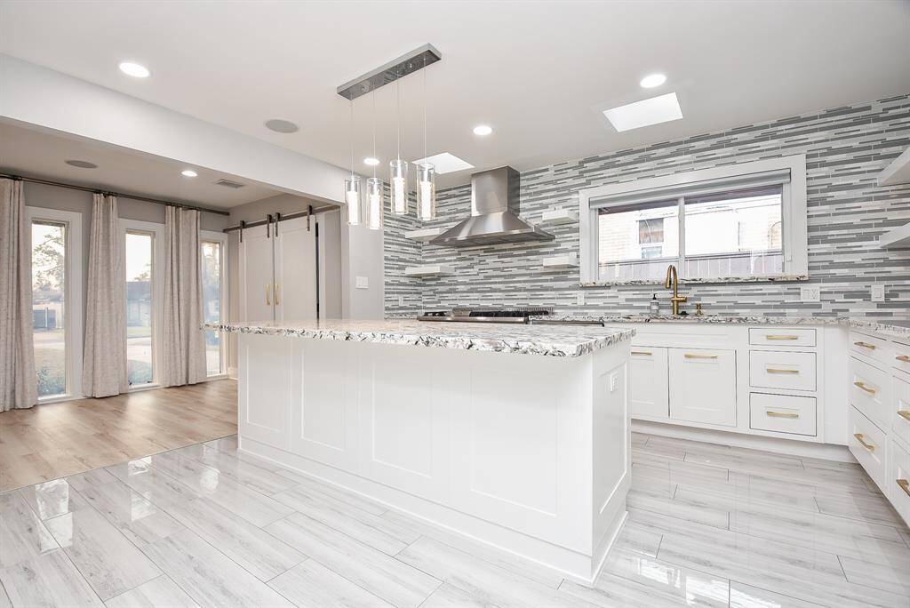 This modern kitchen features sleek white cabinetry with gold accents, a large center island with a granite countertop, and stainless steel appliances. The open layout is complemented by a stylish tile backsplash and ample natural light from the large windows.