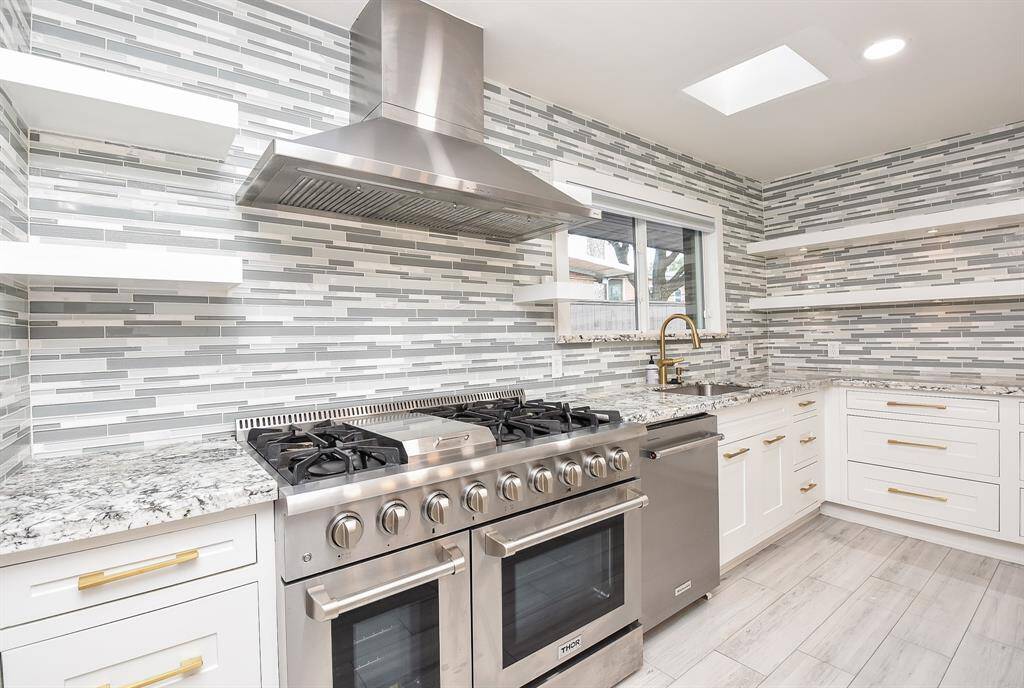 This modern kitchen features sleek stainless steel appliances, a large gas range, and a stylish tile backsplash. It includes ample white cabinetry with gold handles, open shelving, and granite countertops. A skylight and window offer natural light, enhancing the bright and airy feel.