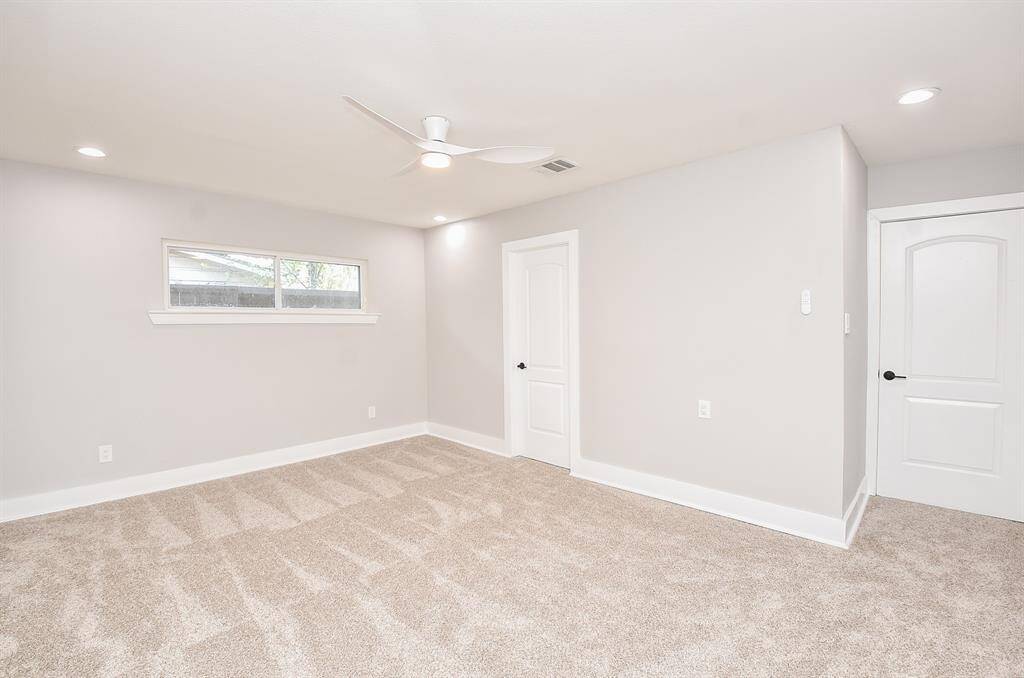 This bright, spacious primary bedroom features neutral carpeting, light gray walls, recessed lighting, and a ceiling fan. It has a window for natural light.