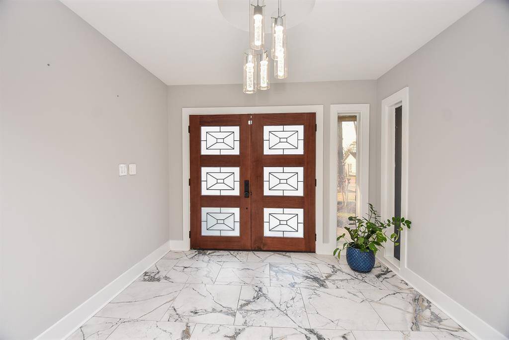 Welcome to a stylish entryway with modern double doors featuring geometric glass, elegant marble floors, and a sleek light fixture. Neutral gray walls and a potted plant create an inviting atmosphere.