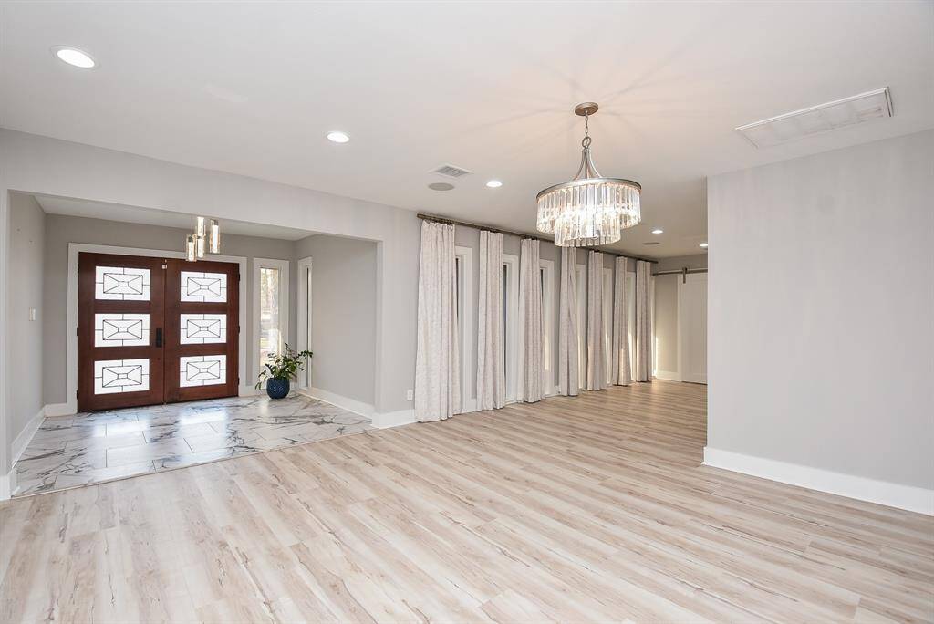 Bright and spacious entryway with light wood flooring, elegant chandeliers, and a modern wooden front door. The area features large windows with curtains, offering plenty of natural light.
