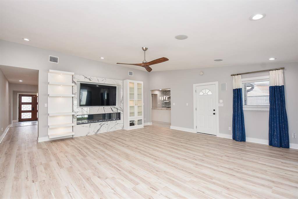 This spacious living room features light wood flooring, a sleek built-in entertainment center with marble accents, and a modern ceiling fan. It includes recessed lighting, bright windows with blue curtains, and entry into the kitchen, creating an inviting and contemporary atmosphere.