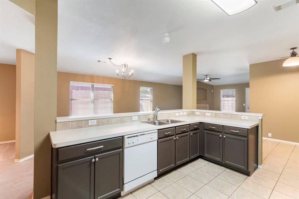 The photo shows a spacious kitchen with an open layout, featuring dark cabinetry, a white countertop, and a built-in dishwasher. The kitchen connects to a bright living area with large windows, neutral walls, and tile flooring, creating a warm and inviting atmosphere.