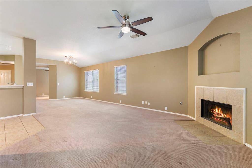This spacious living area features neutral walls, a cozy tiled fireplace, carpeted floors, and a ceiling fan. Large windows allow natural light, and the open layout flows into the kitchen, creating a welcoming atmosphere for gatherings.