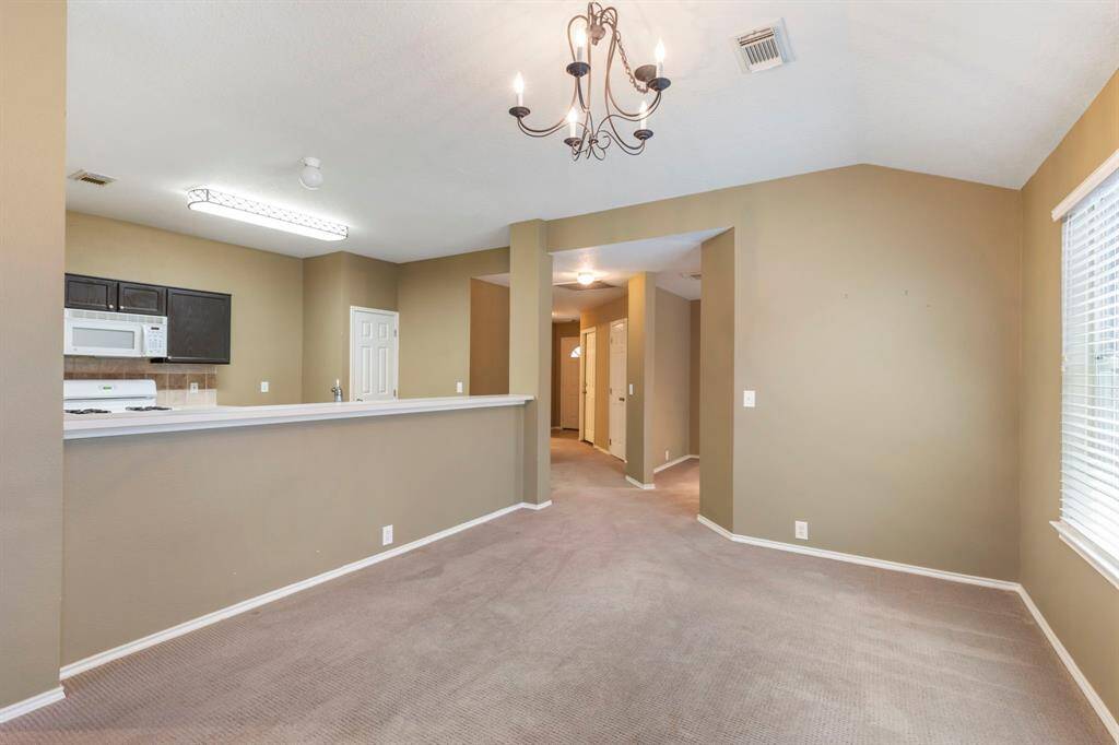 This photo shows an open-concept living space with a view of a kitchen and dining area. The kitchen features dark cabinetry, a white stove, and microwave. The room is carpeted and painted in neutral tones, with a chandelier and ample natural light from a window.