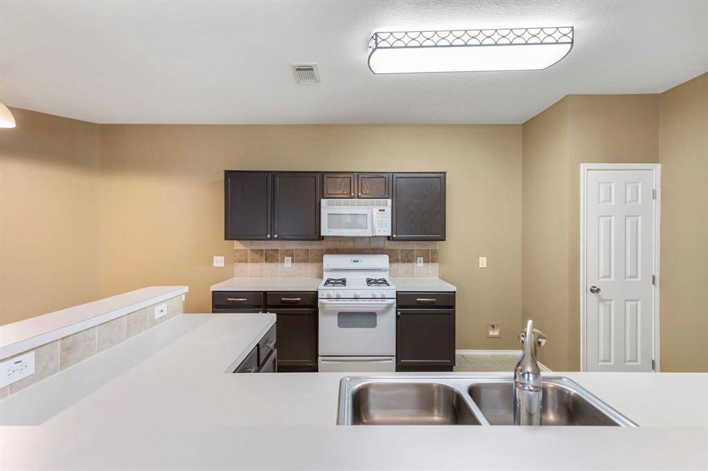 This kitchen features a modern design with dark cabinets, stainless steel double sink, and white appliances, including a gas stove and microwave. The neutral walls and ample lighting create a bright, inviting space.