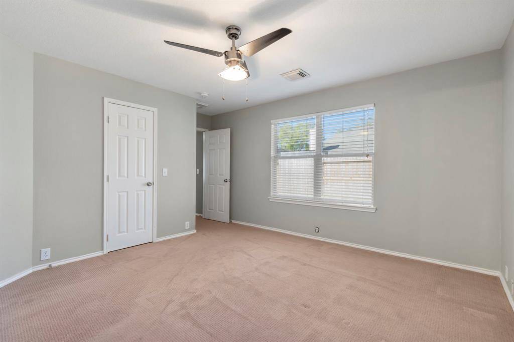 This is a spacious, carpeted bedroom with neutral-colored walls, featuring a large window for natural light and a ceiling fan for comfort. It has a standard closet and an adjacent door.