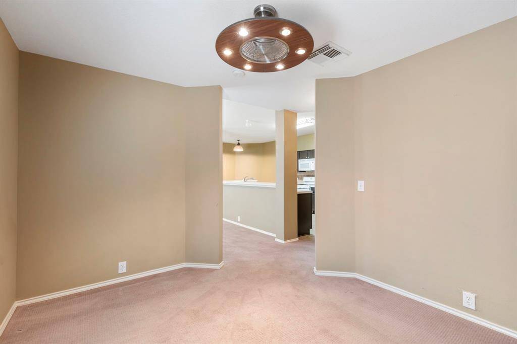 This room features neutral beige walls and carpet, with a modern ceiling light fixture. It opens into a kitchen area with white cabinetry and appliances, creating a cozy and connected living space.