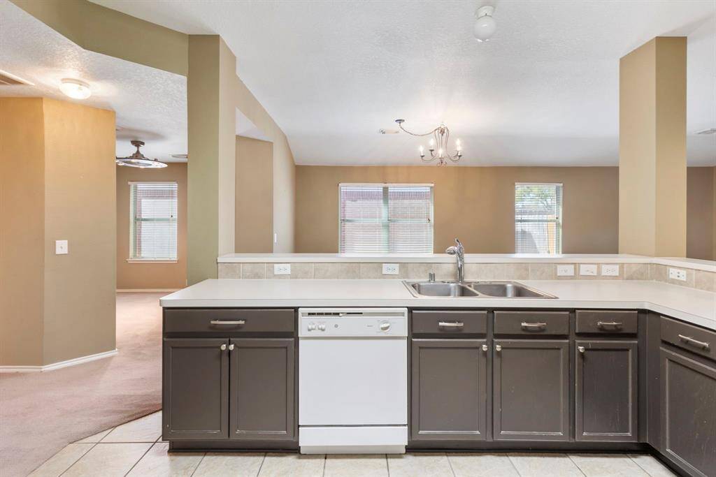 This photo shows a spacious, open-concept kitchen with dark cabinetry, a dual sink, and a dishwasher. The kitchen island overlooks a bright living area with large windows and a ceiling fan, offering a seamless flow for entertaining.