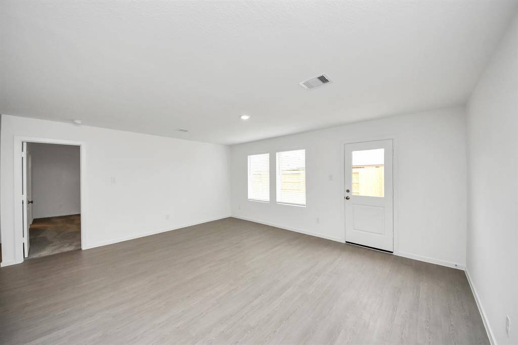 Bright and spacious living area with light wood flooring, white walls, and ample natural light from two windows. There's an adjacent room leading to the primary bedroom and a door leading outside.