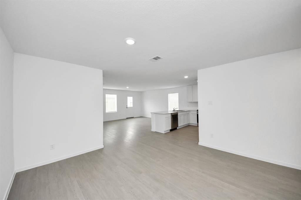 This is a spacious, open-concept living area with light-colored flooring and white walls. It flows into a modern kitchen featuring a large island and stainless steel appliances. Natural light enters through multiple windows, enhancing the bright ambiance.
