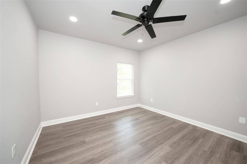 This cozy bedroom featuring a modern ceiling fan, gray wood-like flooring, crisp white walls, and a window for natural light. Perfect for personalizing to your taste and enjoy a relaxing day.