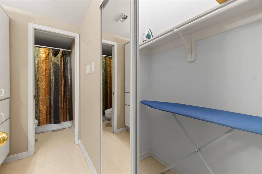 compact laundry or utility area with an ironing board and shelf space, adjacent to a bathroom with a colorful shower curtain. The mirror on the wall, adding depth to the area.