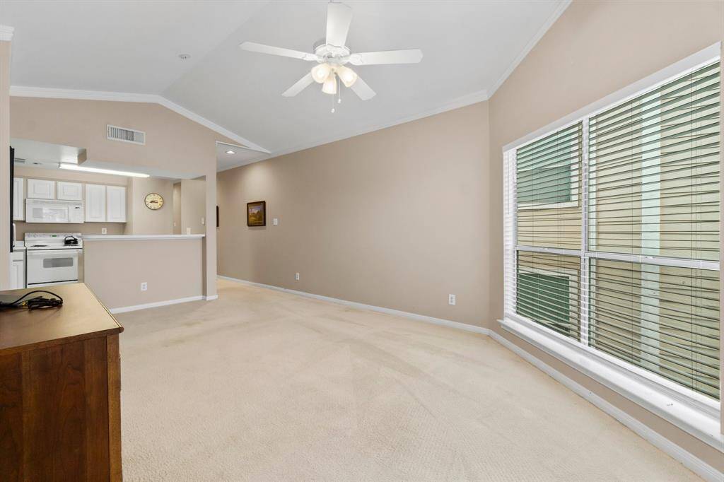 bright, open living space with neutral beige walls and carpet. It features a large window with blinds, a ceiling fan, and an adjoining kitchen with white cabinets and appliances. The area is well-lit and has a cozy, inviting feel.