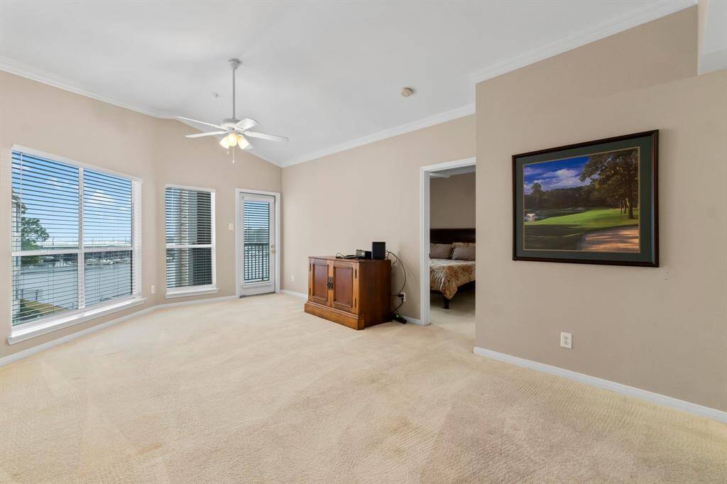Spacious living room with large windows offering a scenic view of the water. Features a ceiling fan, neutral walls, and carpet flooring. There's a door leading to a balcony and an entrance to a bedroom. Perfect for enjoying natural light and serene surroundings.