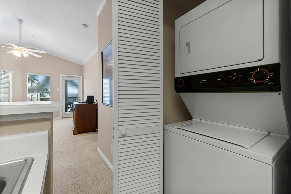 compact laundry area with a stacked washer and dryer, adjacent to an open living space featuring neutral walls, carpet flooring, and large windows with blinds.