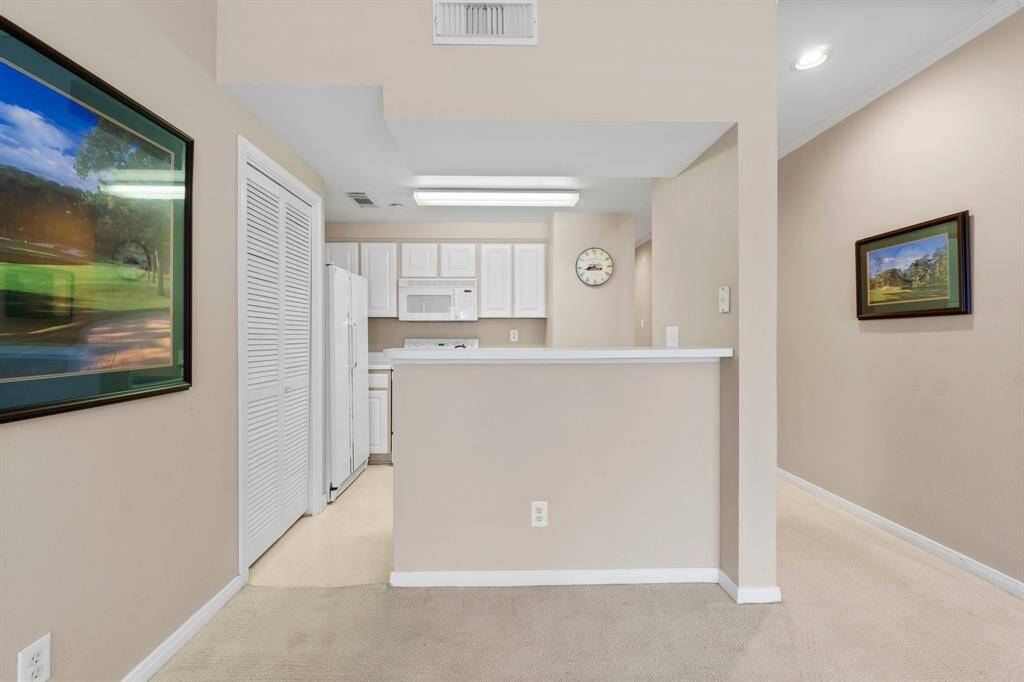 a cozy kitchen area with white cabinetry and appliances, open to a small dining counter. The walls are a neutral color, complemented by framed artwork. The space appears well-lit and inviting.