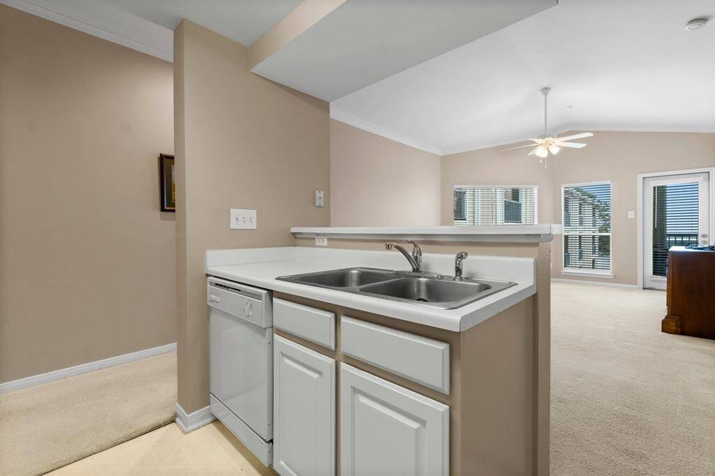 open kitchen with a white dual sink and dishwasher, overlooking a spacious living area. The living room has carpet flooring, large windows for natural light, a ceiling fan, and access to a balcony or patio through glass doors, creating a bright and airy atmosphere.
