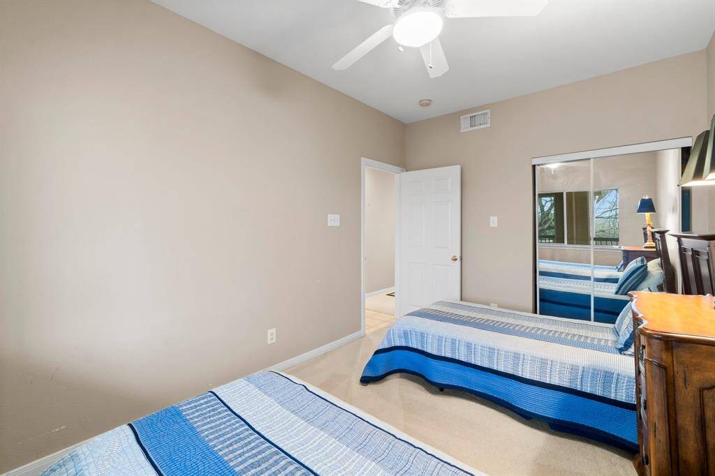 cozy bedroom with neutral walls and carpet, featuring a ceiling fan. There are two twin beds with blue bedding, a wooden dresser, and a mirrored closet door that reflects natural light, creating a bright and inviting atmosphere.