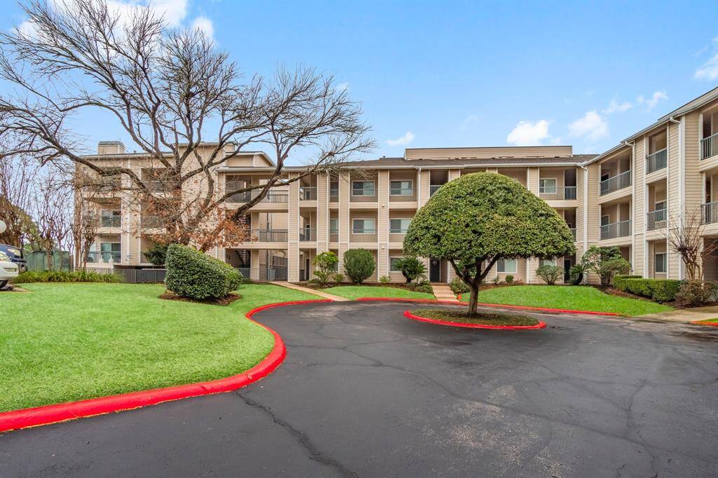 three-story complex with a clean, well-maintained exterior. The building is surrounded by tidy landscaping, including trimmed shrubs and a distinctive, neatly-pruned tree in the center of a circular driveway. The area is well-kept, ideal for potential buyers seeking a pleasant living environment.