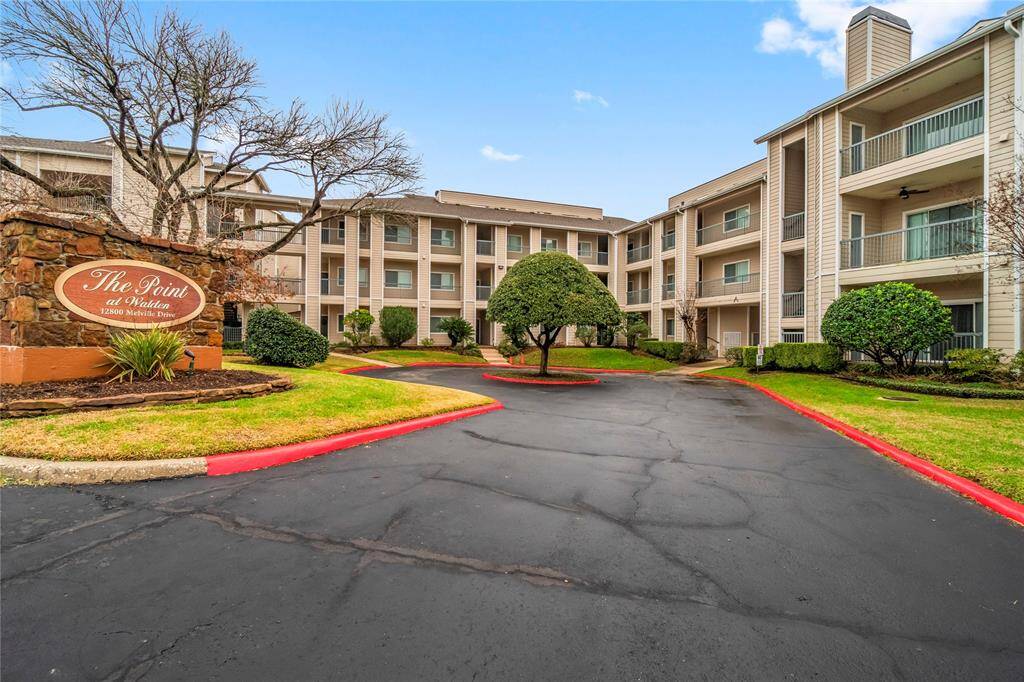 well-maintained  complex with three-story buildings, balconies, and manicured landscaping. The entrance features a welcoming sign, and there's ample parking space with clearly marked driveways.
