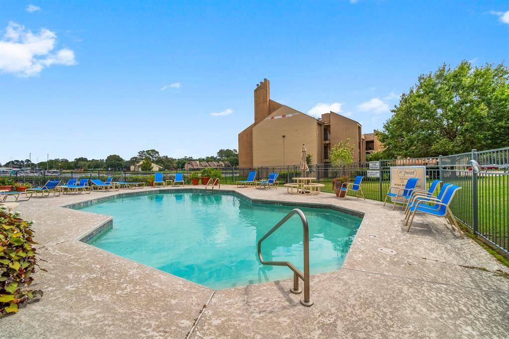 fenced community pool with lounge chairs and tables, set in a landscaped area with trees and a building in the background, perfect for relaxation and outdoor enjoyment.