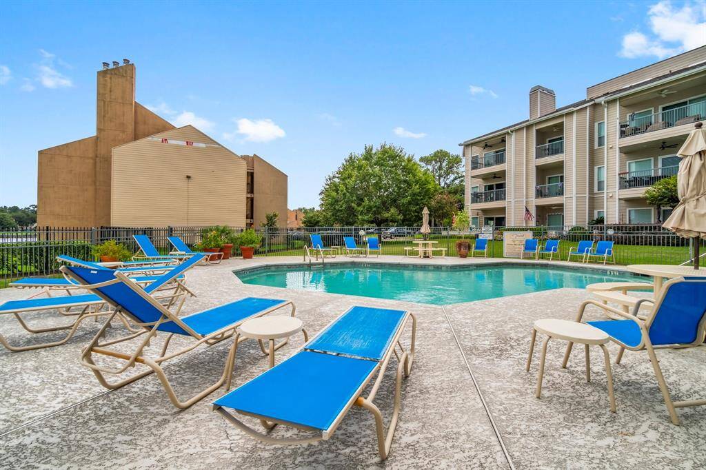 residential community pool area with blue lounge chairs and umbrellas, surrounded by well-maintained landscaping and adjacent to multi-story buildings. It's a relaxing outdoor space perfect for leisure and gatherings.